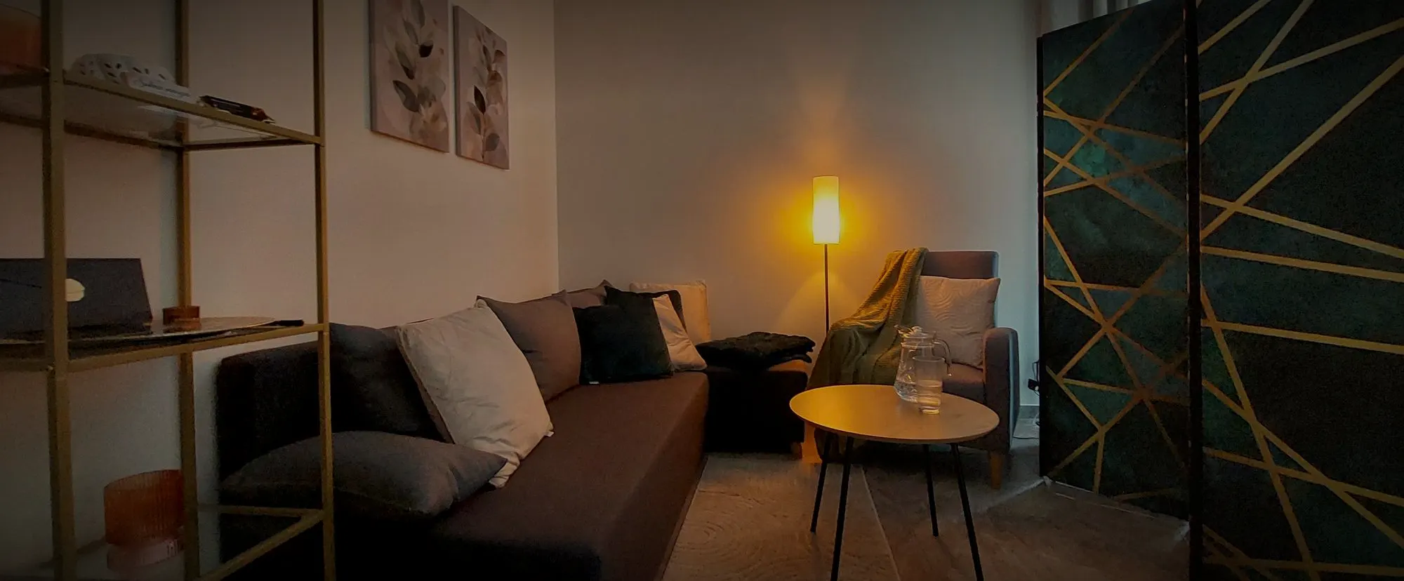 Kącik wypoczynkowy: sofa z poduszkami, okrągły stolik i zapalona lampa obok złoto-zielonego parawanu